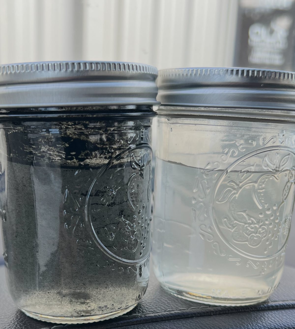 Two mason jars side by side on a truck dashboard: the left jar holds black solids-laden produced water taken upstream of the strainer, the right jar holds clear filtered water taken downstream